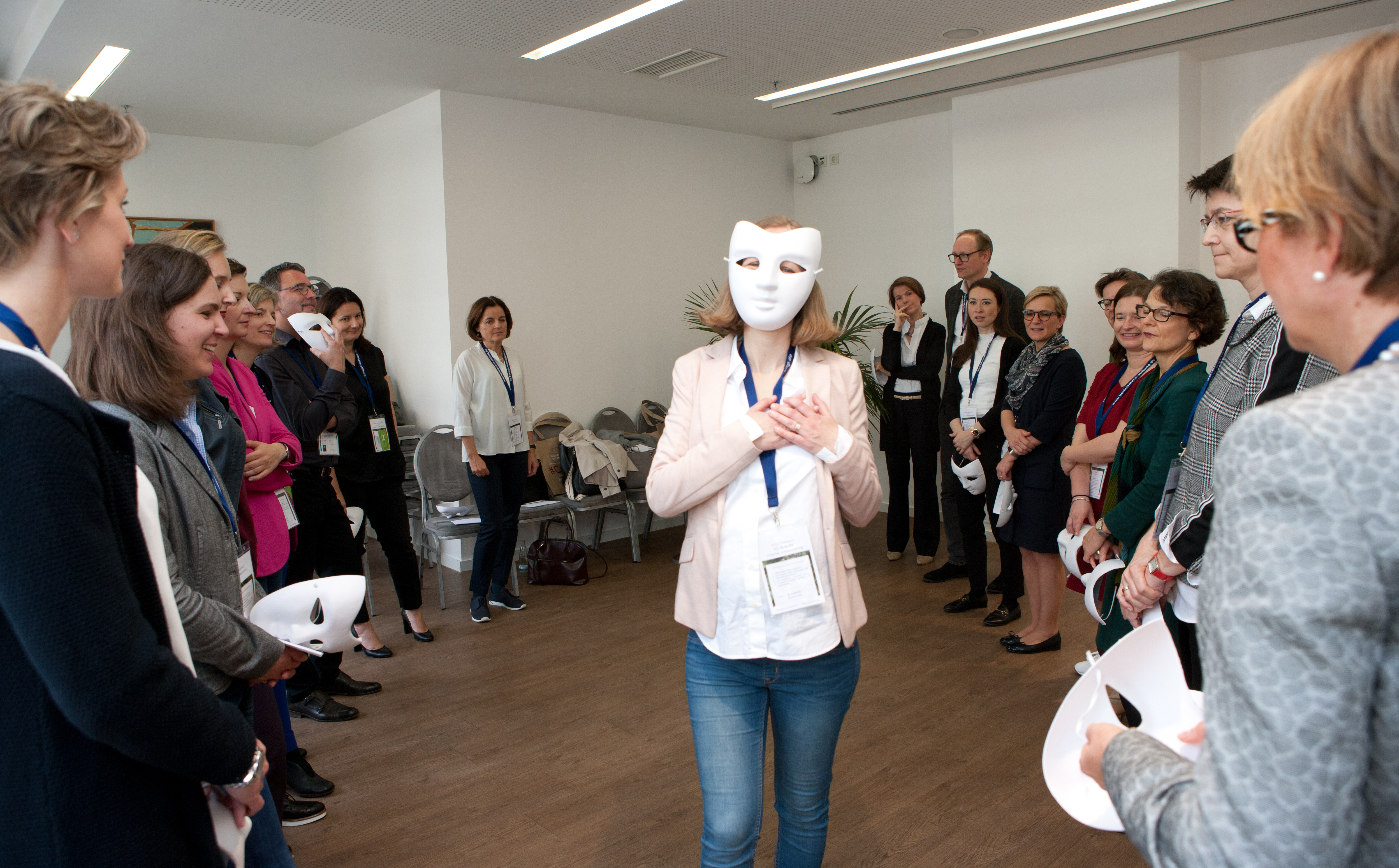 eine Person mit einer weißen Maske vor dem Gesicht, darum im Halbkreis weitere Personen