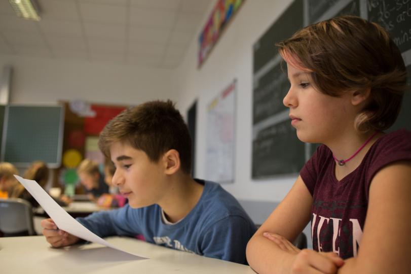 Ein etwas 11jähriger Schüler liest etwas von einem Blatt ab, neben ihm sitzt eine Schülerin